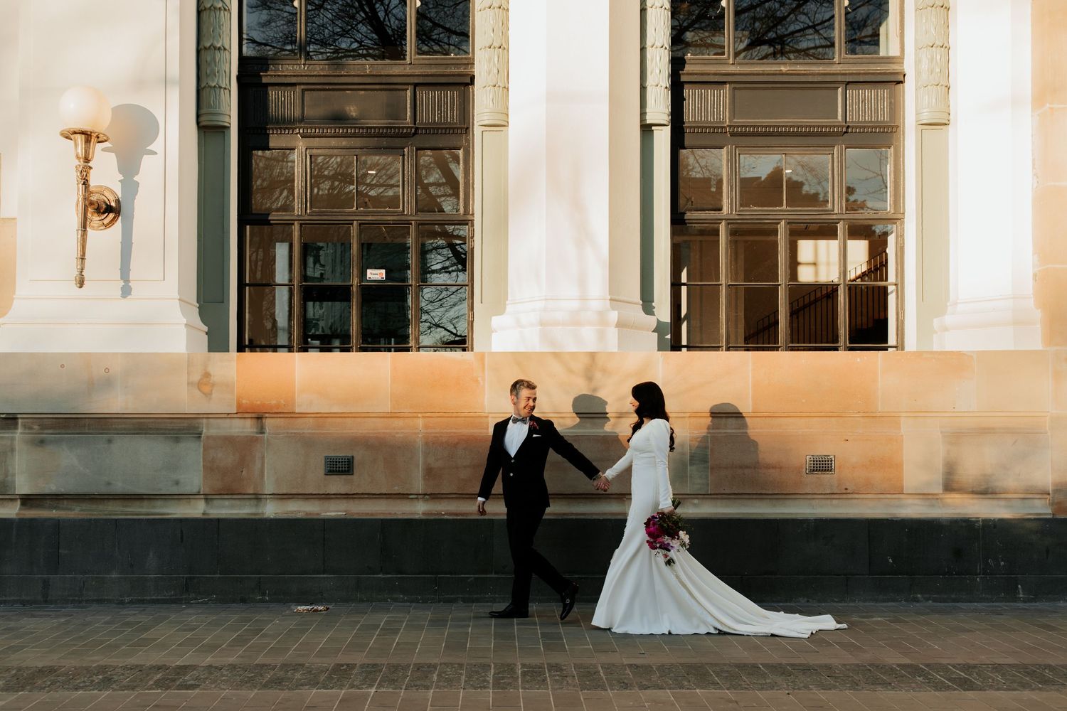 Elegant wedding couple poses outside historic building with dramatic lighting.