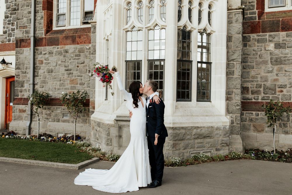 Wedding couple poses against stone church wall with ornate gothic windows.