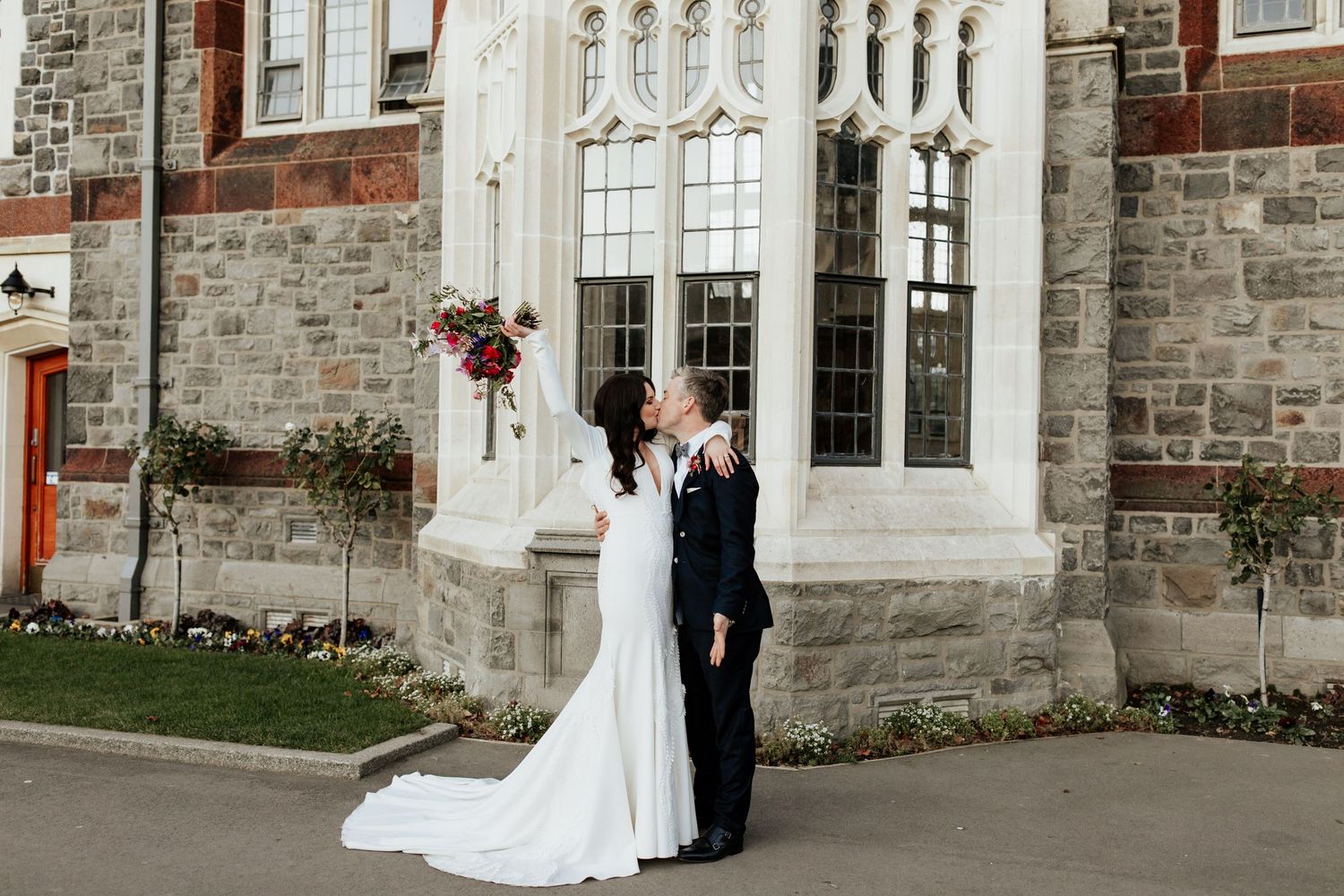 Wedding couple poses against stone church wall with ornate gothic windows.