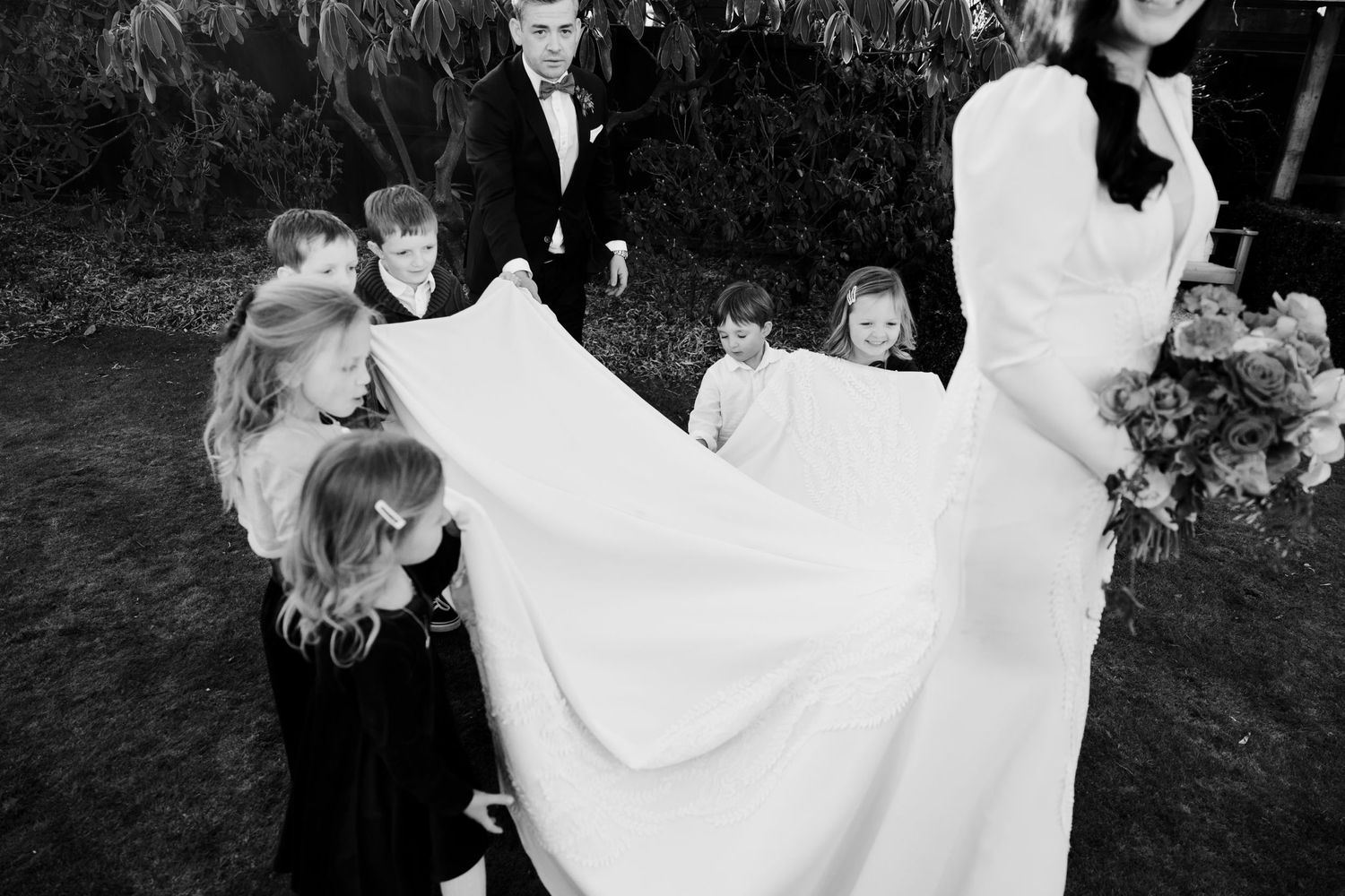 A black and white photo captures children playing with a flowing wedding dress train outdoors.