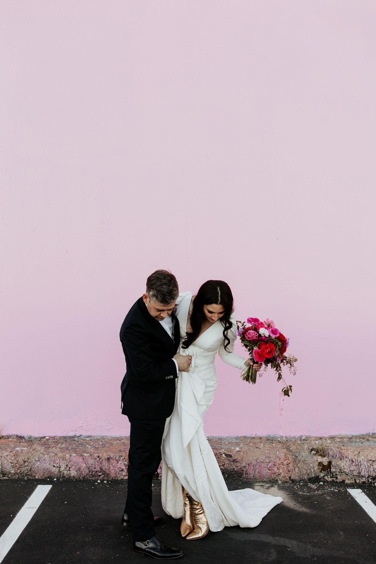 Wedding couple shares intimate moment against pink wall with vibrant red and pink bouquet in parking lot.