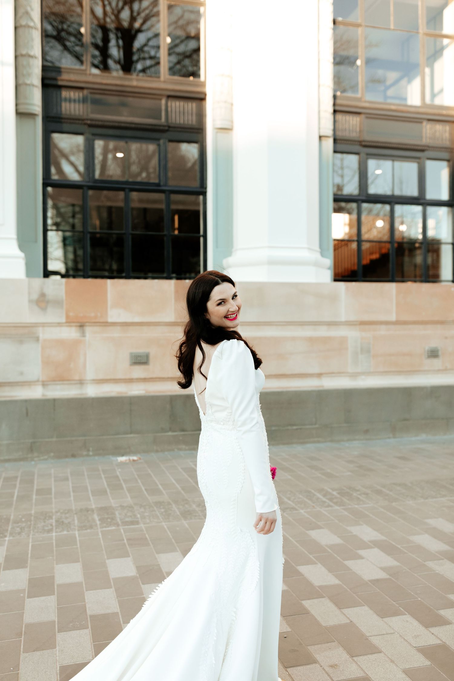 Bride in long-sleeved gown looks over shoulder in artistic architectural portrait.