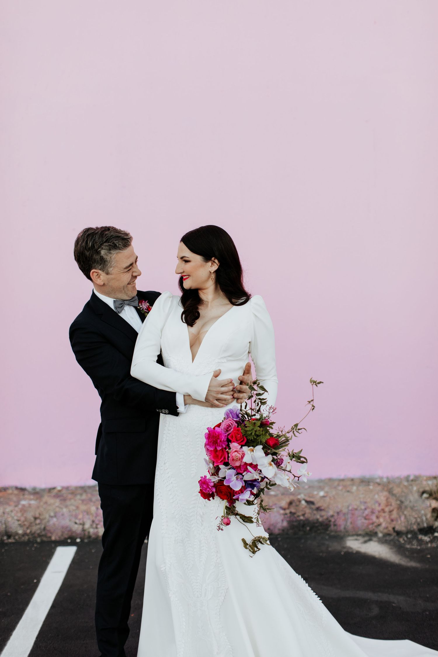Elegant wedding couple embracing against pink wall with bright floral bouquet.