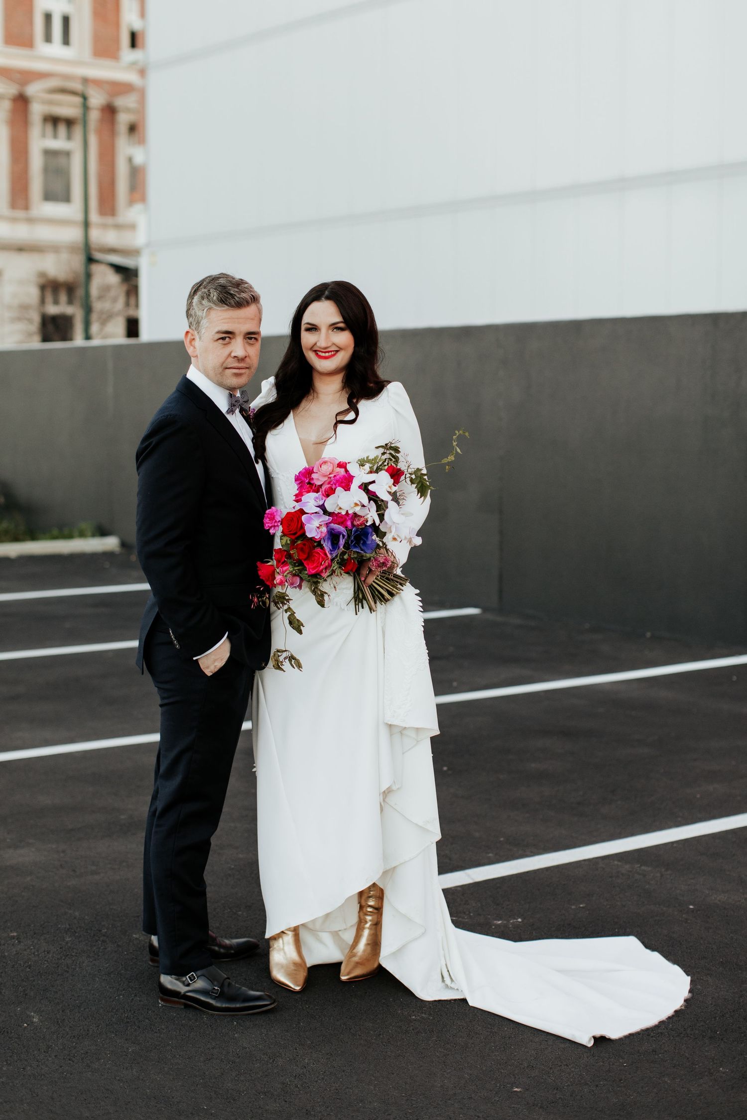Modern wedding portrait in urban setting with dramatic white gown and colorful bouquet.