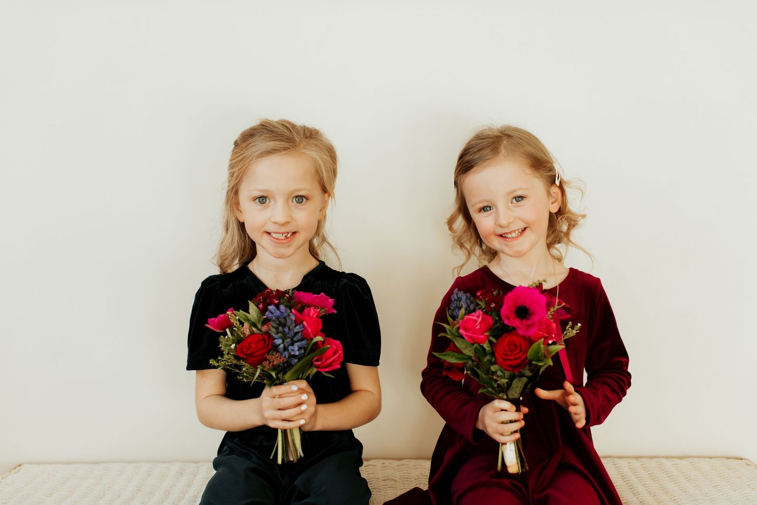 Two young girls in formal dresses holding red rose bouquets against white background.