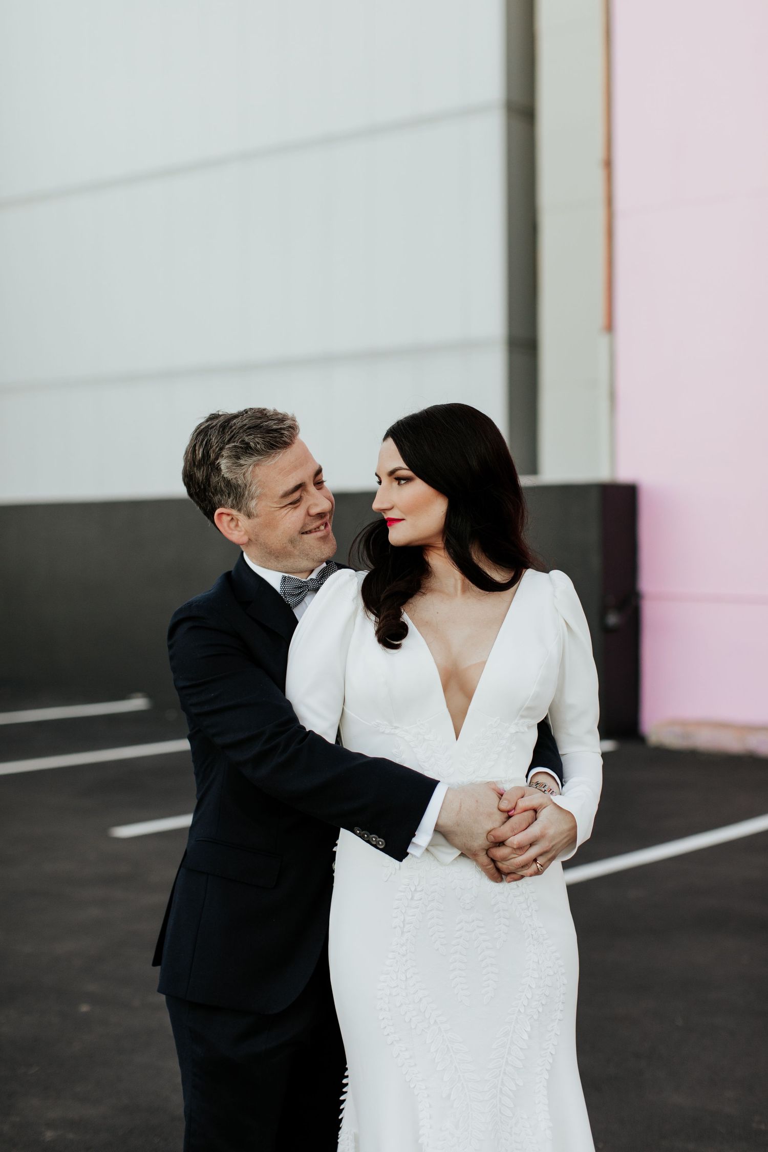 Romantic couple embraces in modern urban setting with pink wall accent.