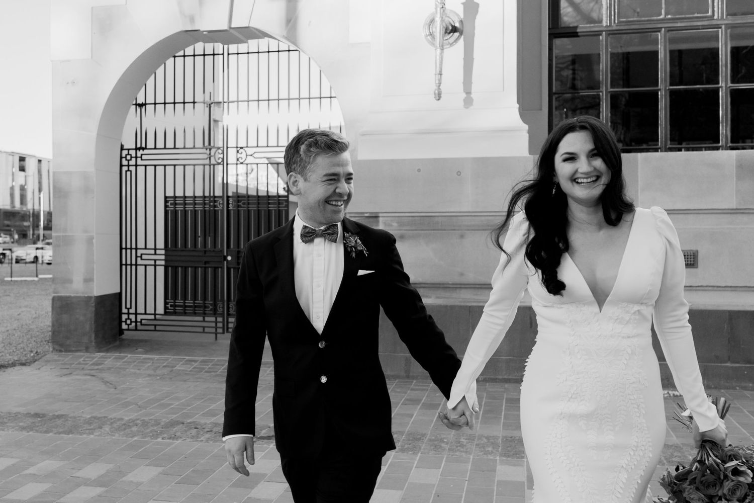 Black and white photo of couple walking hand in hand past wrought iron gate.