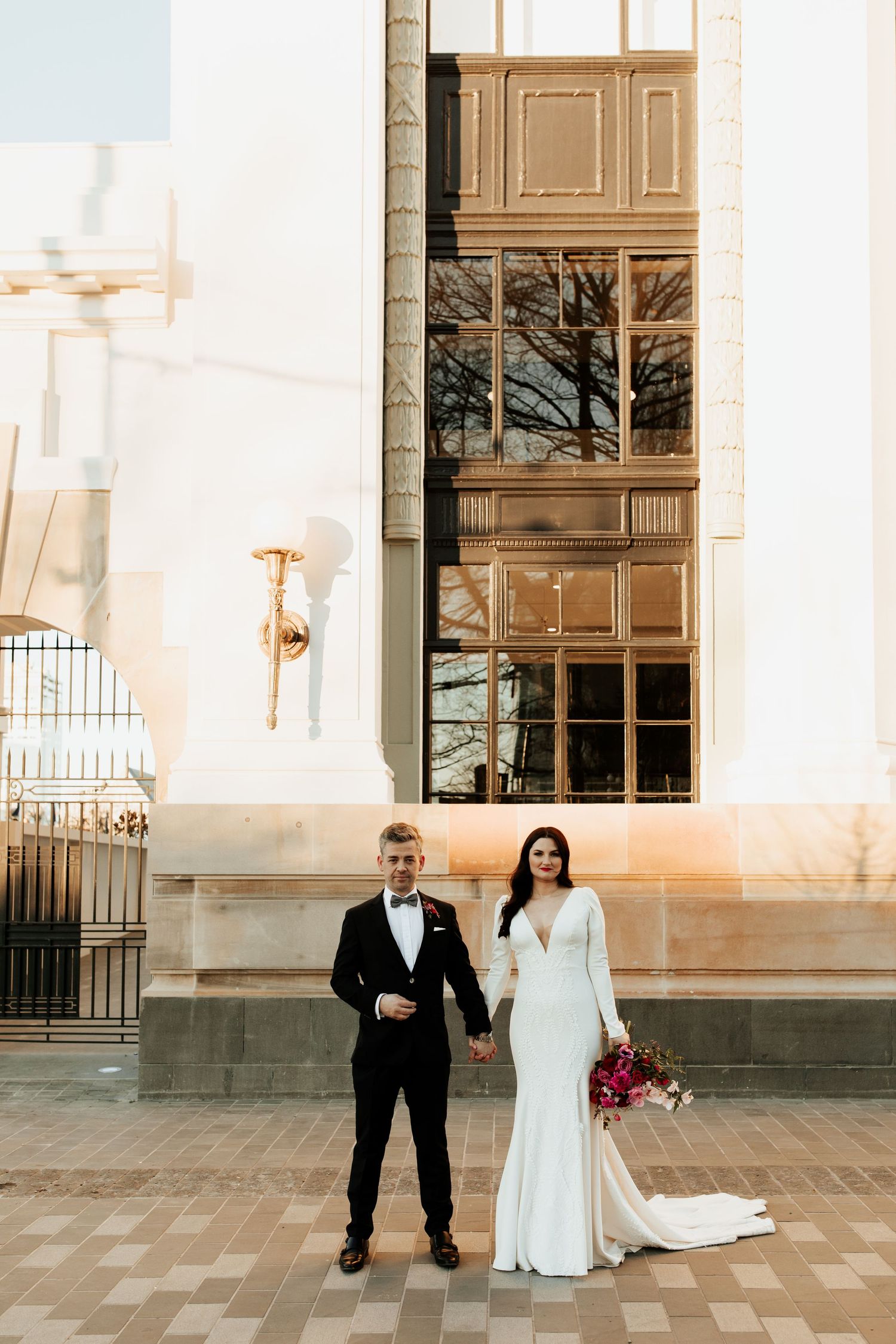 Elegant wedding couple standing in front of historic building with tall windows and brick pavers.