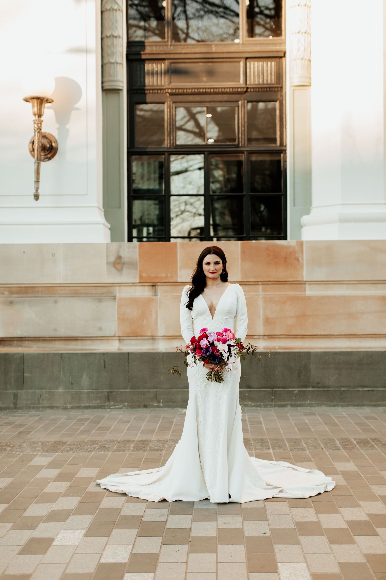Bride in flowing white gown holding colorful bouquet against classical building facade.