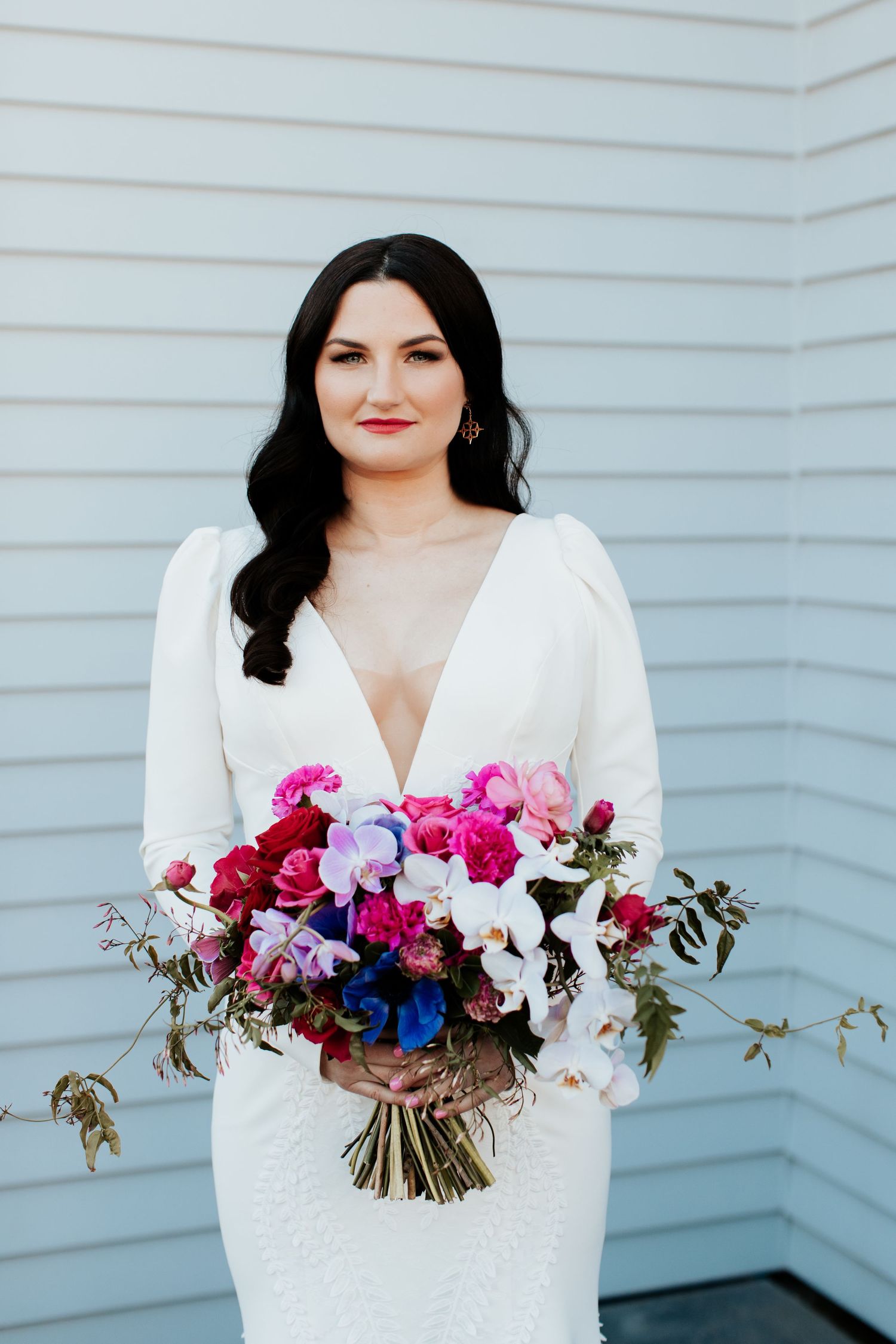 Elegant bridal portrait featuring long-sleeve white dress and colorful bouquet against grey siding.