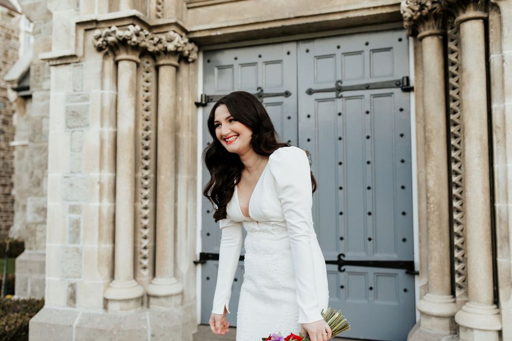 An elegant bride in a fitted white gown holds pink flowers against Gothic architecture.