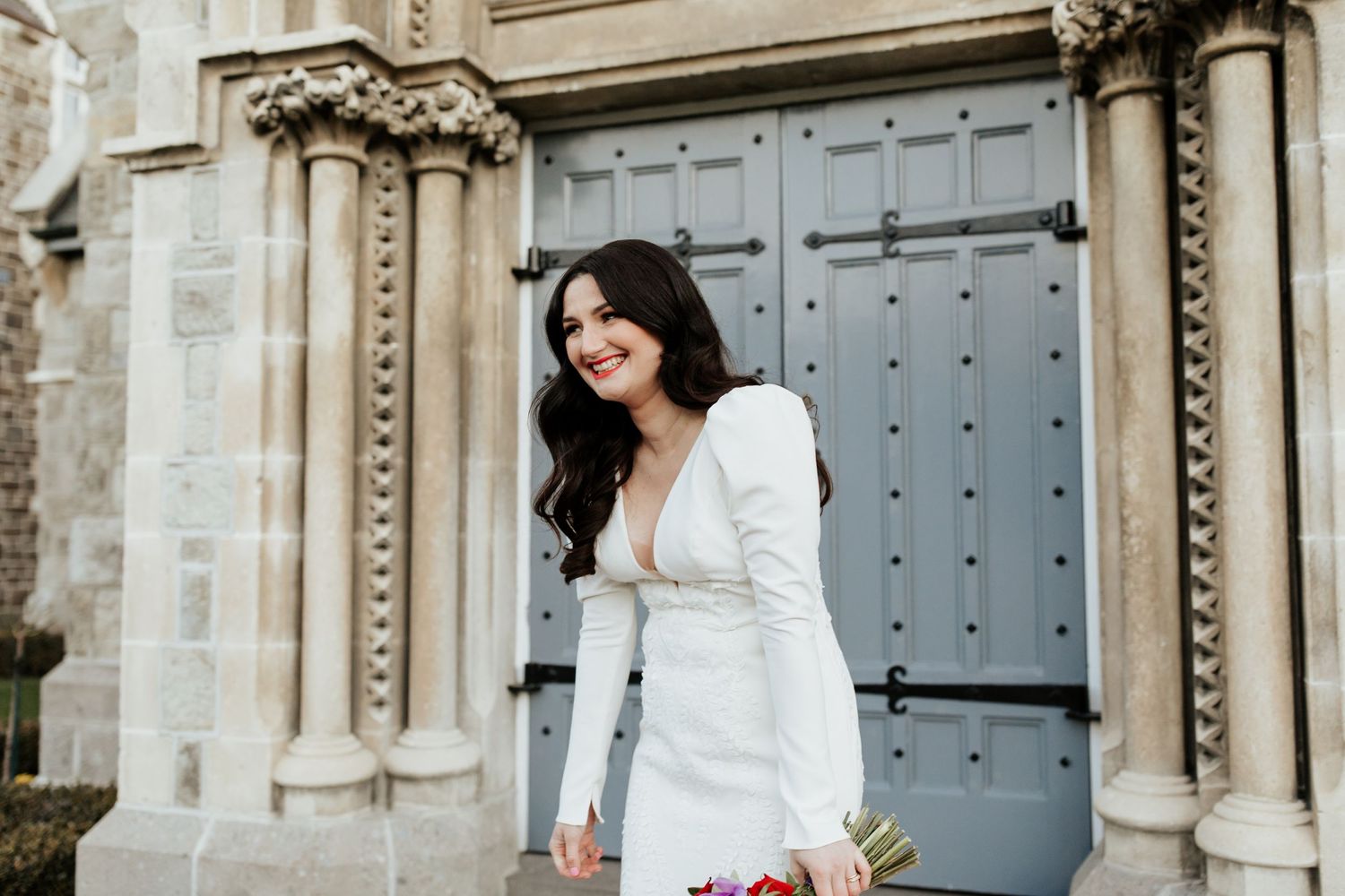 An elegant bride in a fitted white gown holds pink flowers against Gothic architecture.