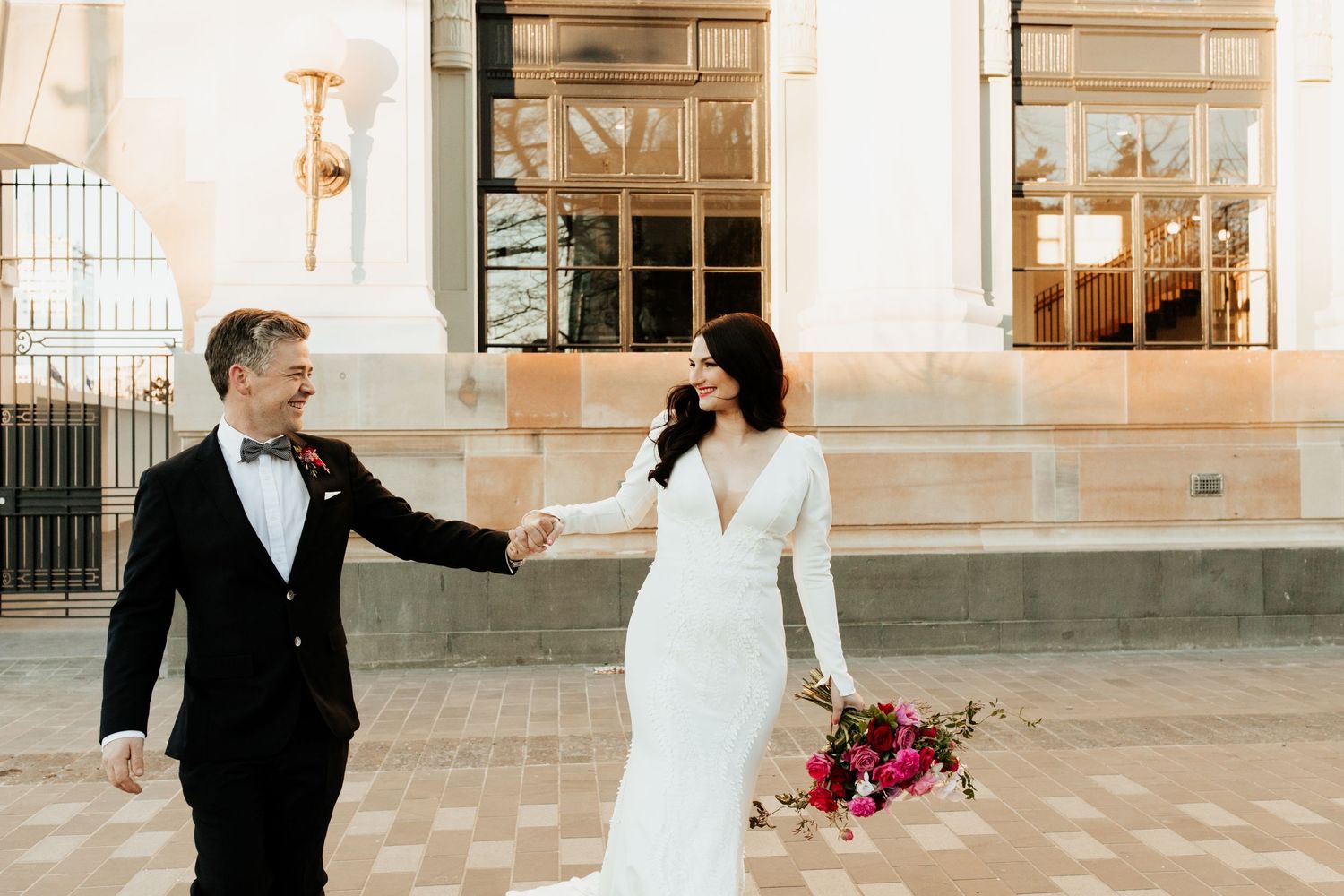 Wedding couple holding hands and walking along stone building with warm sunlight.