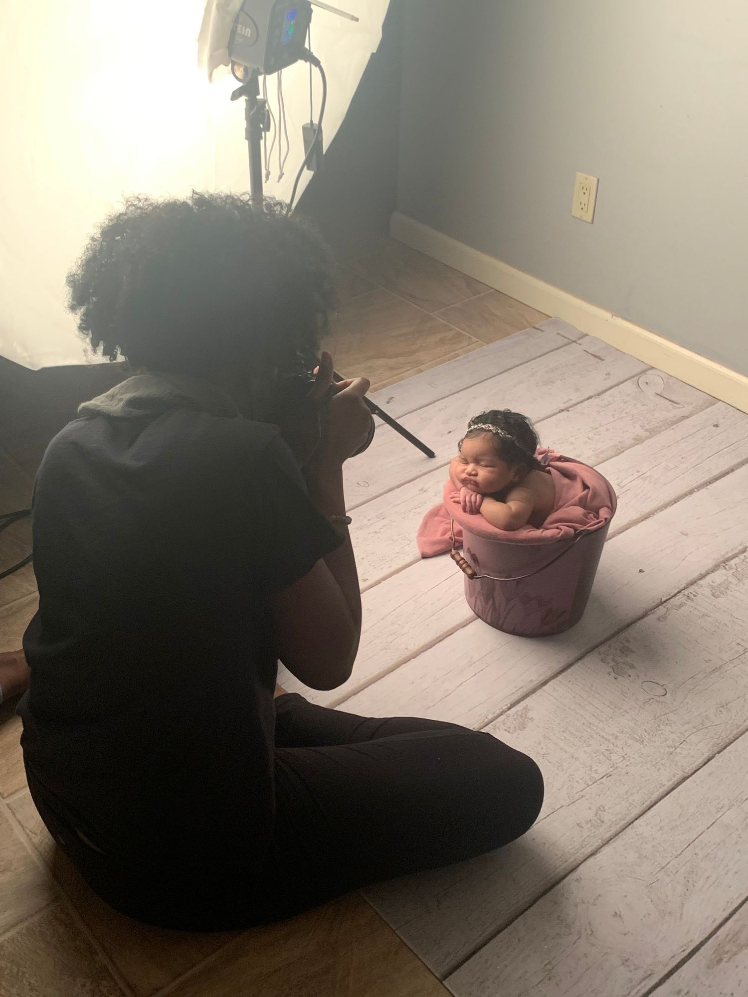 Behind-the-scenes photo of photographer capturing baby in pink bucket on wooden floor.