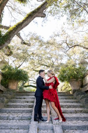 Romantic couple embraces on stone steps under sunlit trees with red dress flowing.