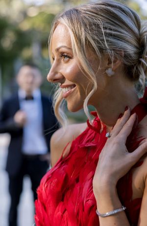 Woman in red feathered dress smiles while touching her neck as figure stands blurred in background.