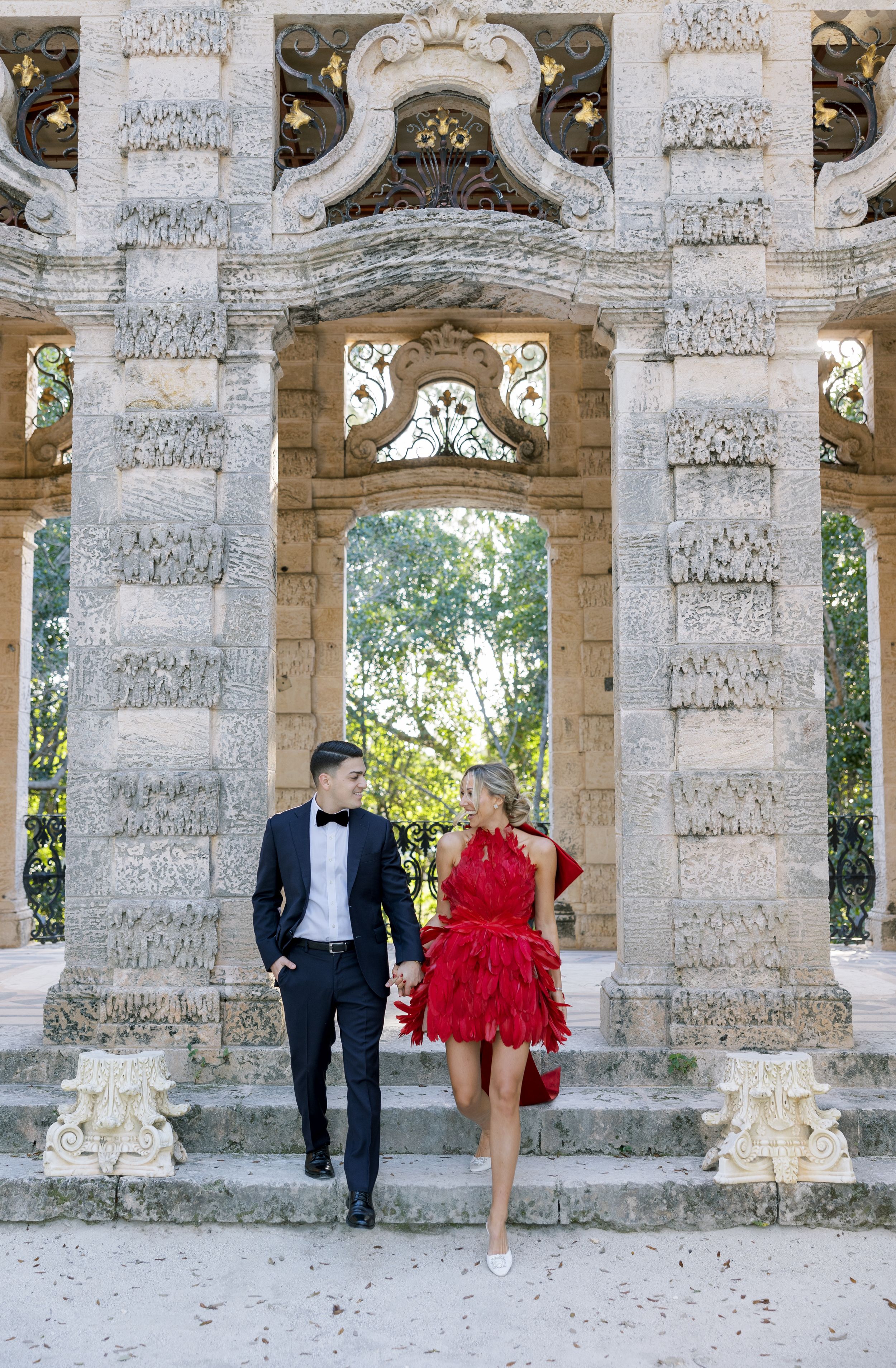A couple poses elegantly in formal attire between ornate stone columns at a historic venue.