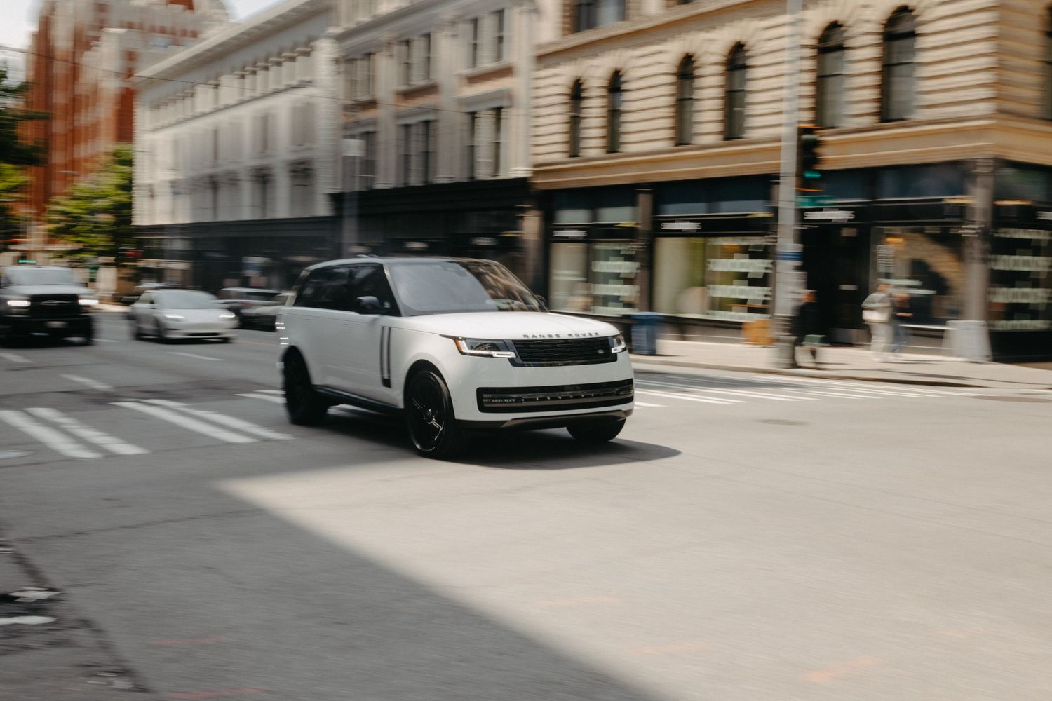 A white Range Rover SUV drives through a city street with motion blur and historic buildings in the background.