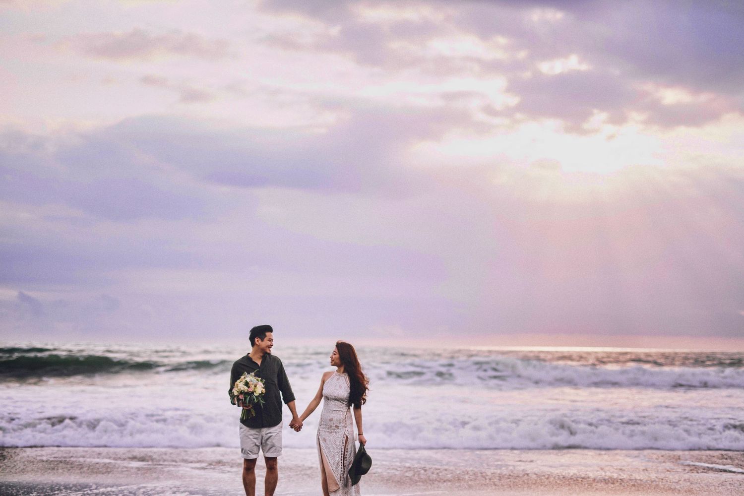 Romantic couple embracing on a scenic beach at sunset with dramatic cloudy skies and crashing waves.