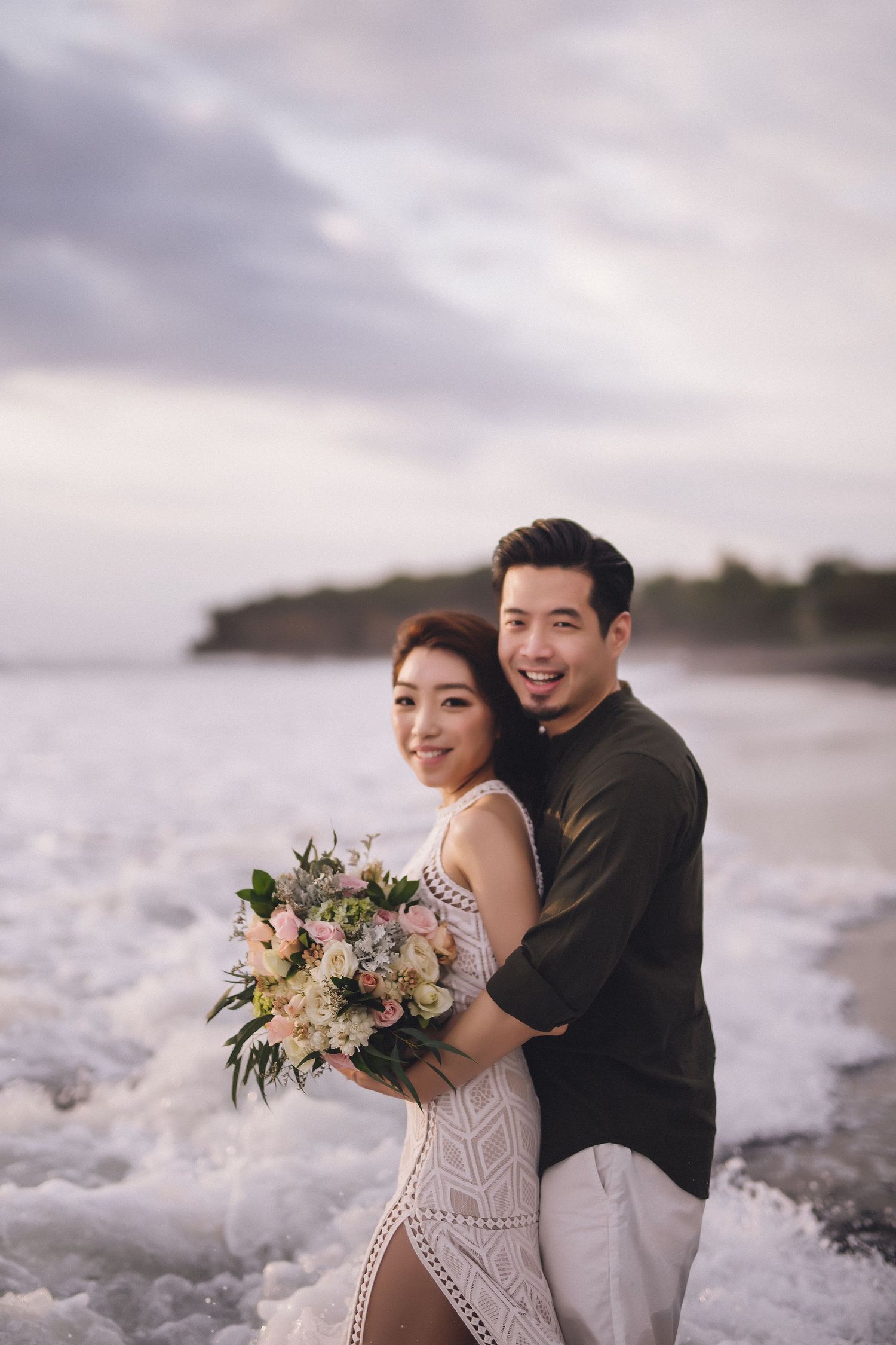 Couple in formal attire share an intimate moment on the beach with a floral bouquet at dusk.