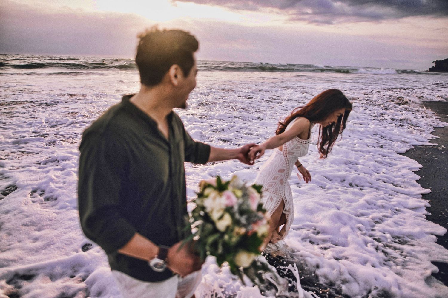 Couple dancing and twirling on the beach with ocean waves and dramatic sky in background.