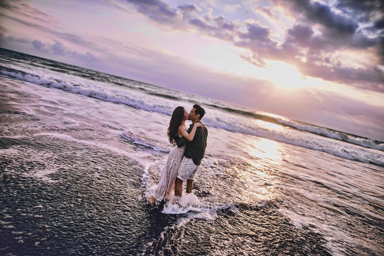 Romantic beach photoshoot with couple silhouetted against stunning sunset and ocean waves.