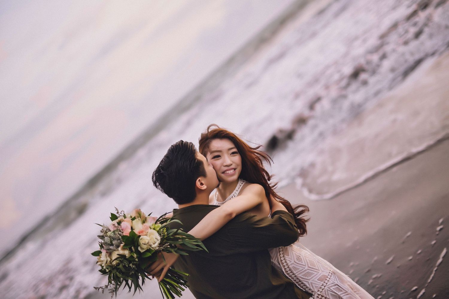 Couple in elegant attire sharing tender moments by the ocean during sunset.