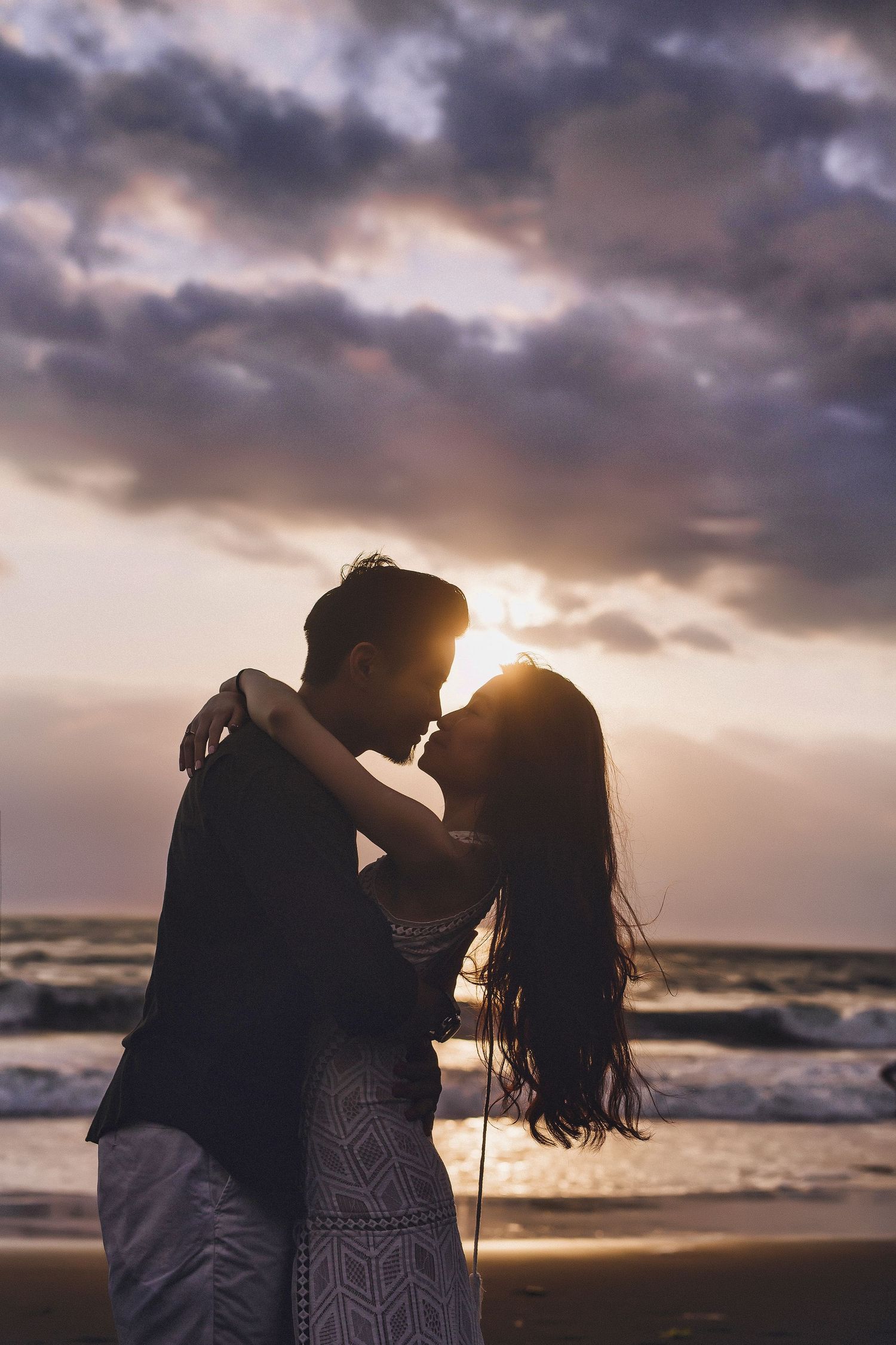 Silhouetted couple embracing on beach with dramatic purple sunset clouds overhead.