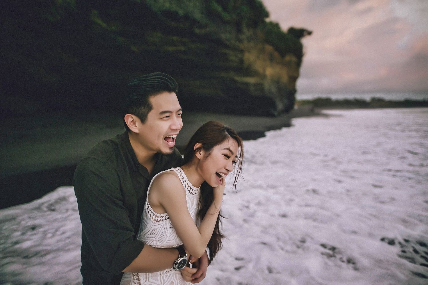 Romantic couple embraces on a dramatic rocky beach coastline during sunset.