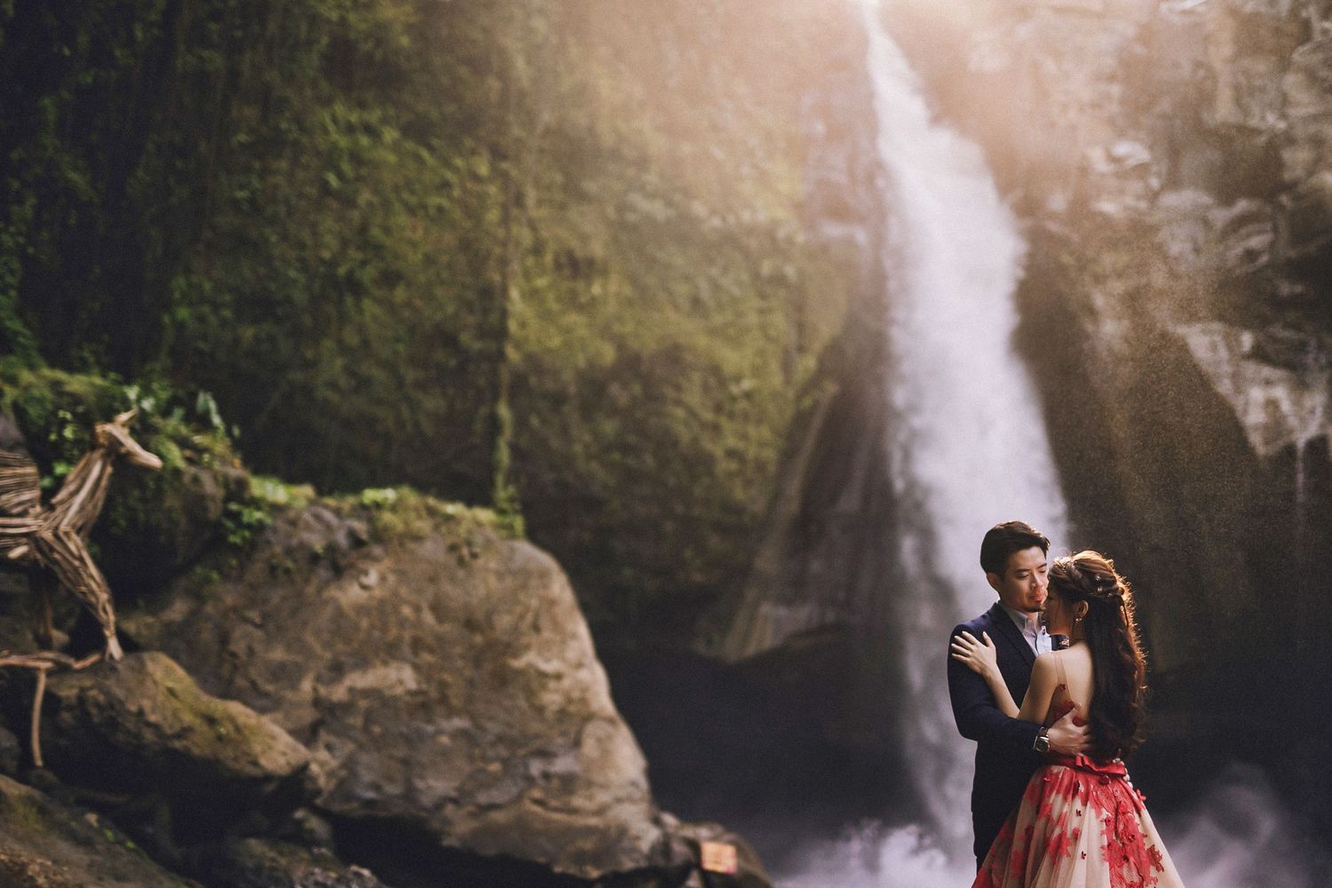 Elegant couple stands before powerful waterfall in formal wear.