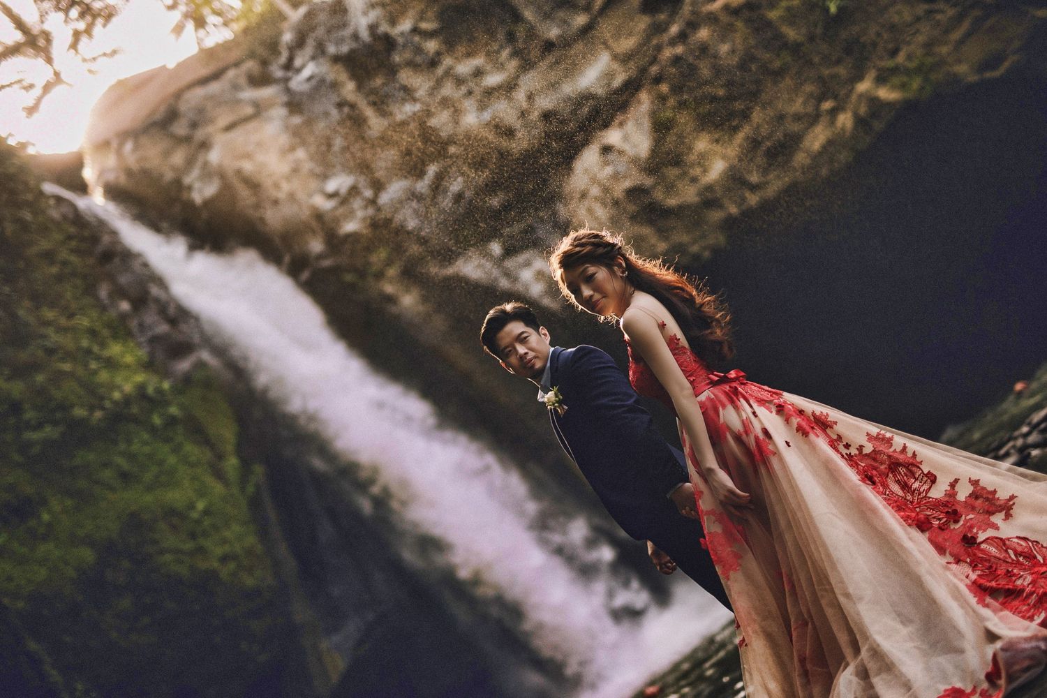 Couple in formal attire embraces near stunning waterfall cascade.
