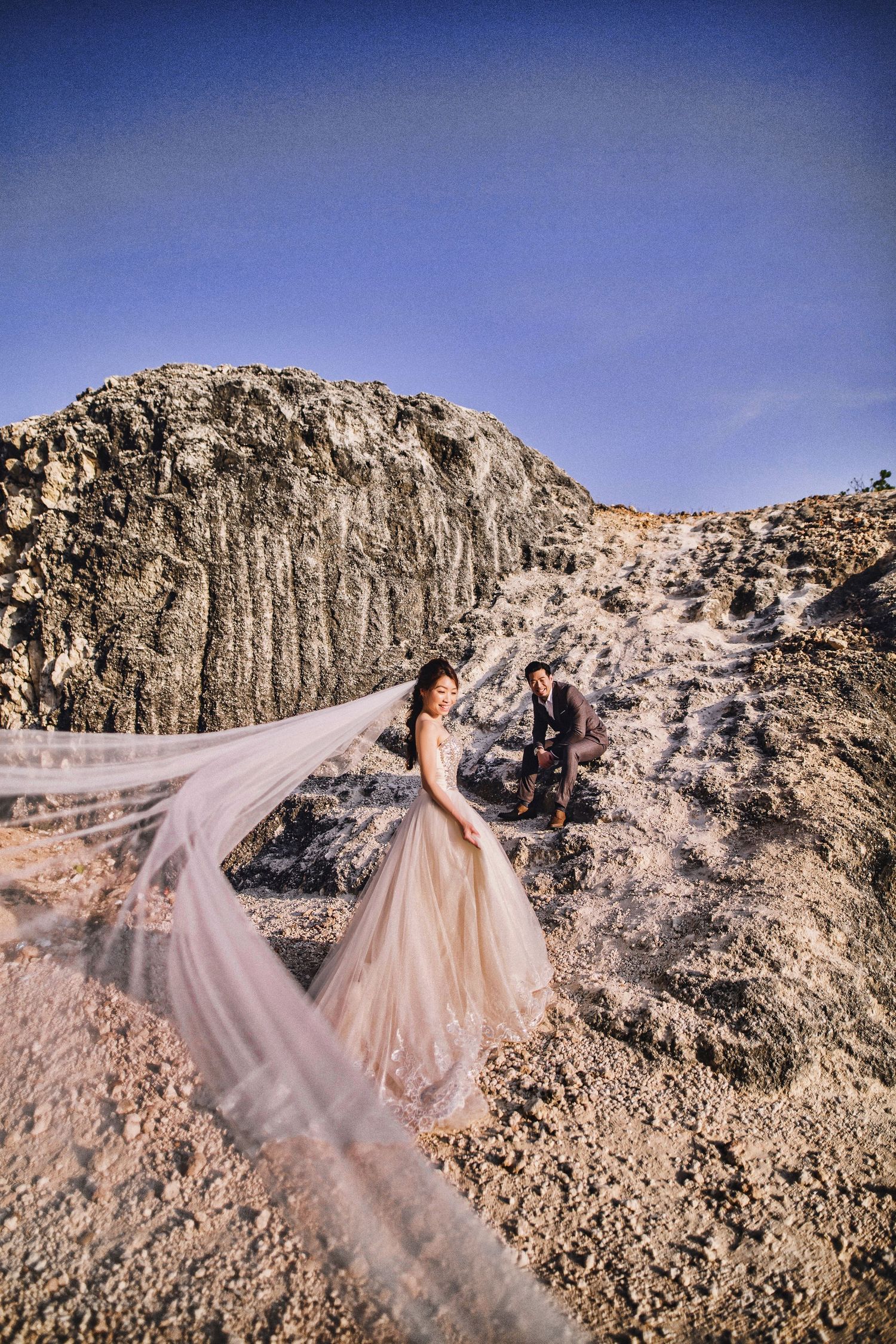 Wedding couple with flowing veil pose on rocky terrain against bright blue sky.