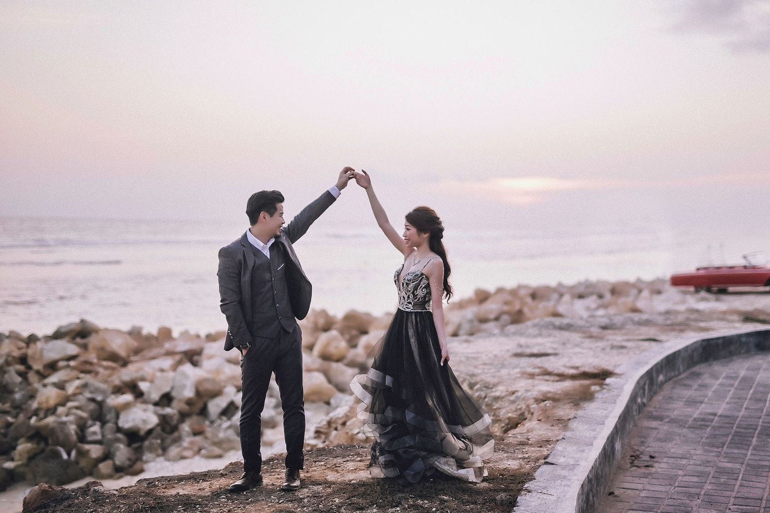 A romantic couple shares an intimate moment on a scenic beach at sunset with dramatic coastal cliffs in the background.