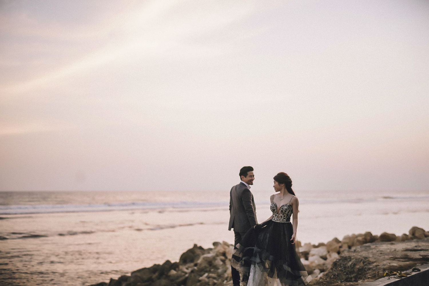 Two people embrace on a rocky beach shoreline during golden hour with waves crashing behind them.