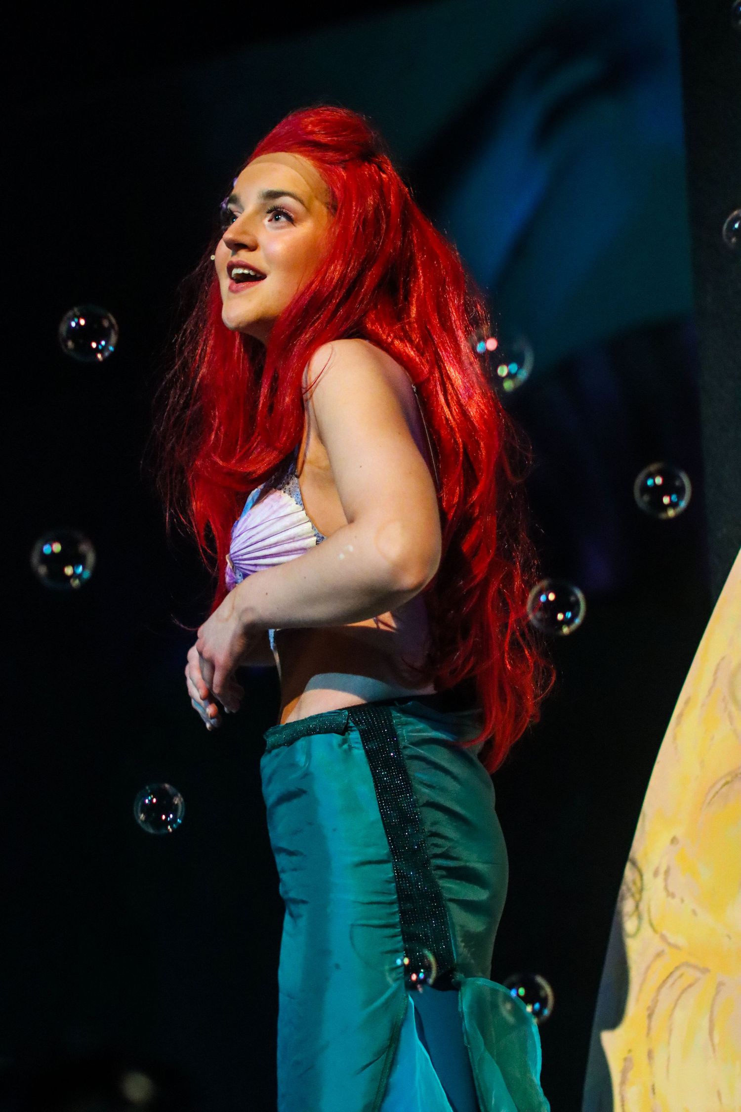 Performer with vibrant red hair in costume with floating bubbles on dark stage.
