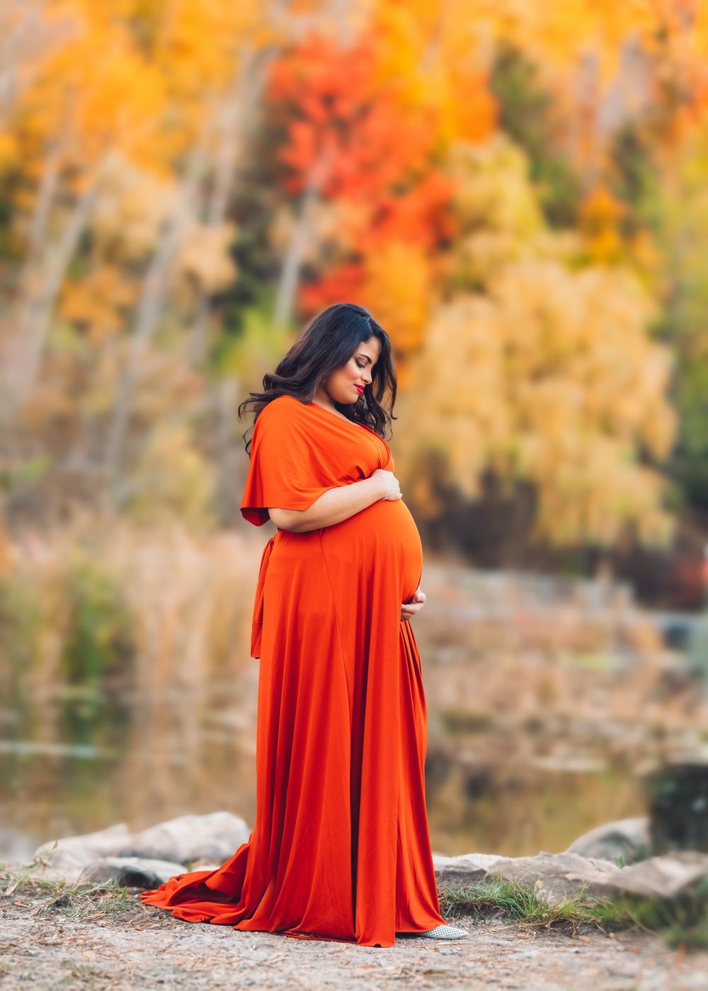 Maternity portrait of an expecting mother in a flowing red gown, standing in a scenic autumn landscape. Toronto maternity photography.