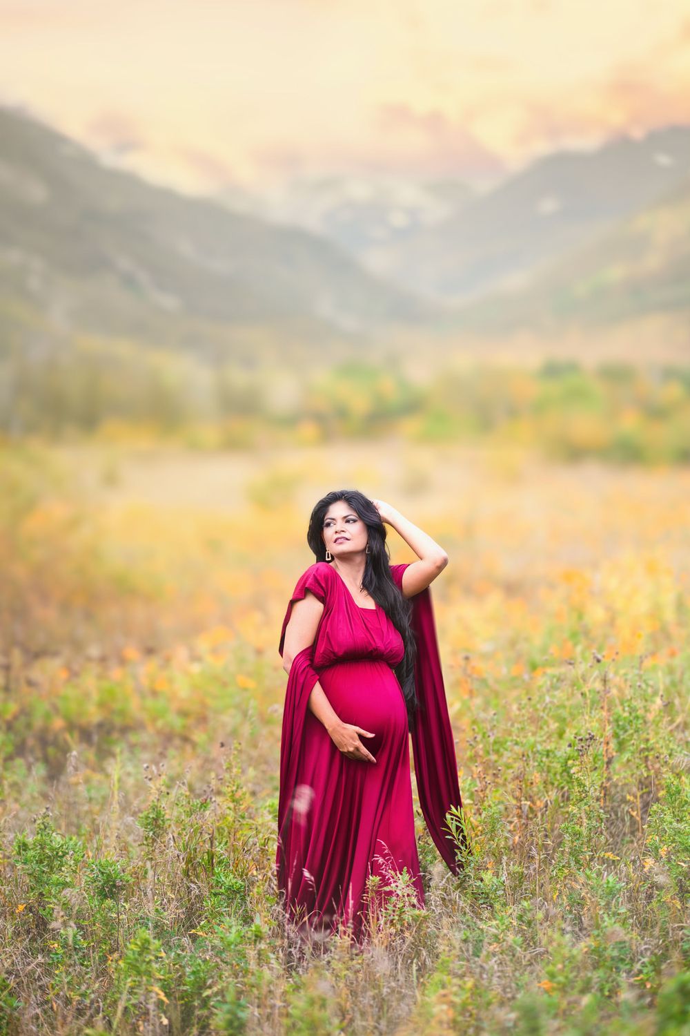 Woman in a flowing red dress standing in a scenic field with mountains in the background. Toronto maternity photography.