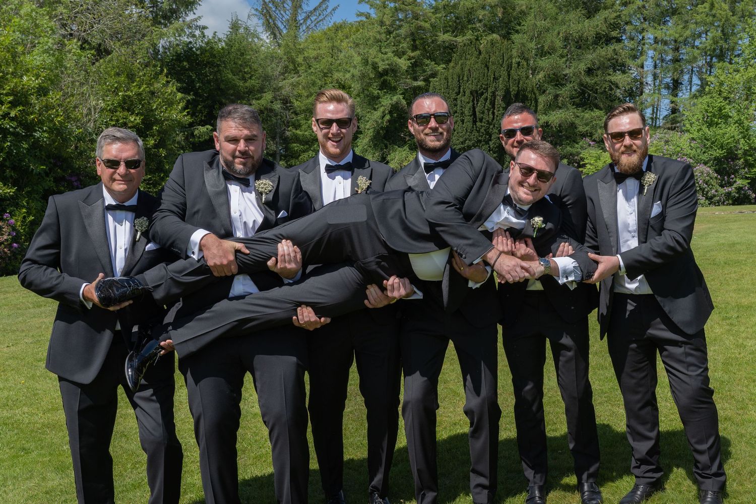 Groomsmen in matching suits playfully lift groom during outdoor photo session.