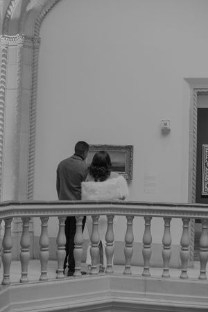 Two people observe artwork from a balcony with white decorative balusters in an art gallery.