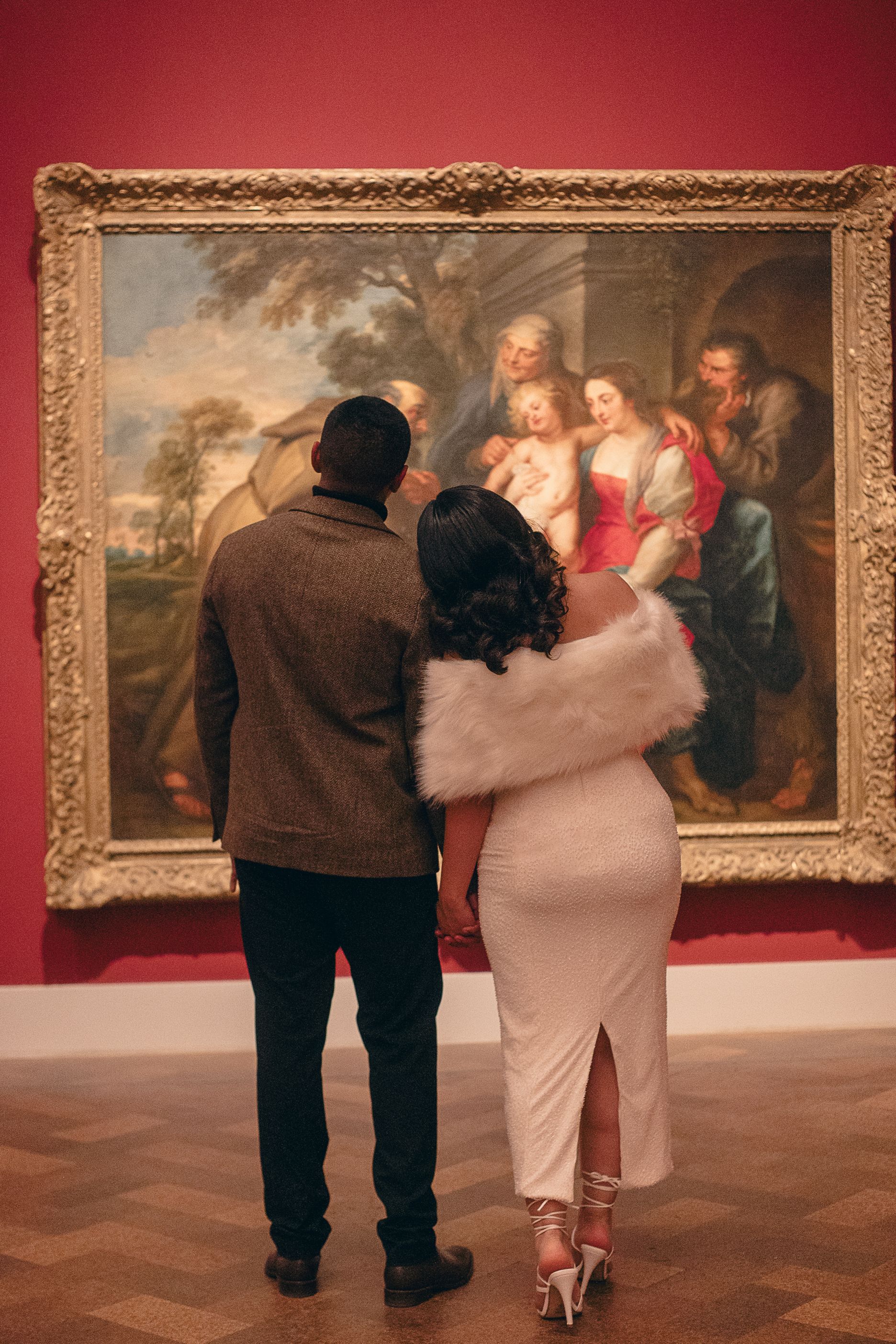 A couple admires artwork together in a museum gallery with red walls and ornate gold frames.