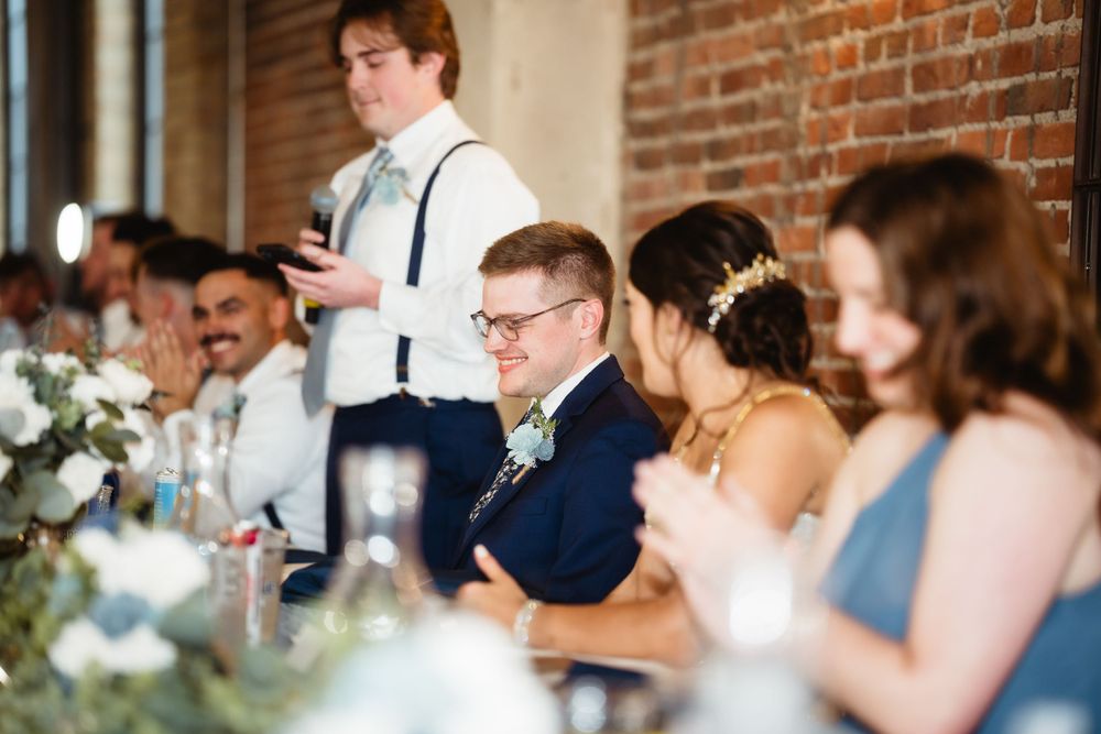 Wedding reception guests seated at long table during speeches in industrial venue.