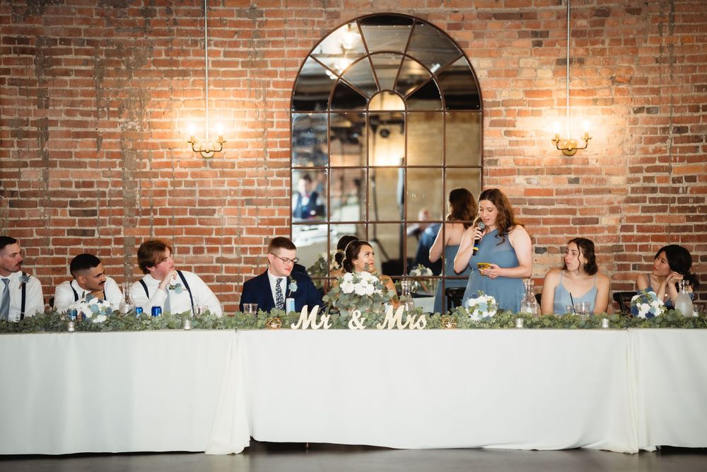 Wedding party seated at head table in venue with brick arch and wall sconces.