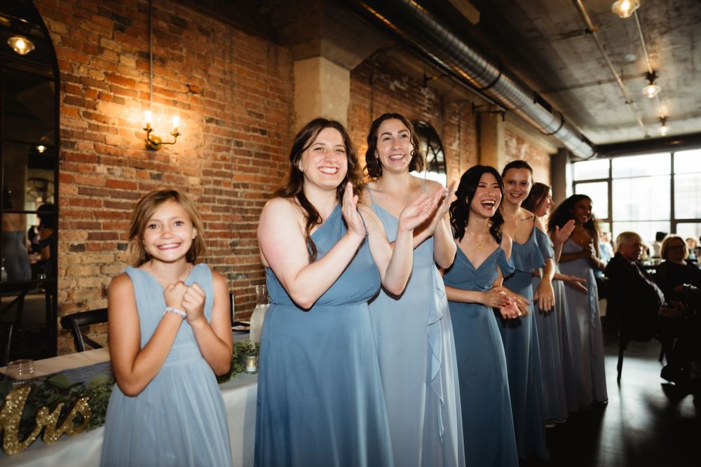 Wedding party in dusty blue dresses clapping and celebrating at indoor venue with exposed brick walls.