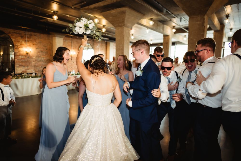 Wedding party dancing and celebrating at reception with brick walls and hanging lights.