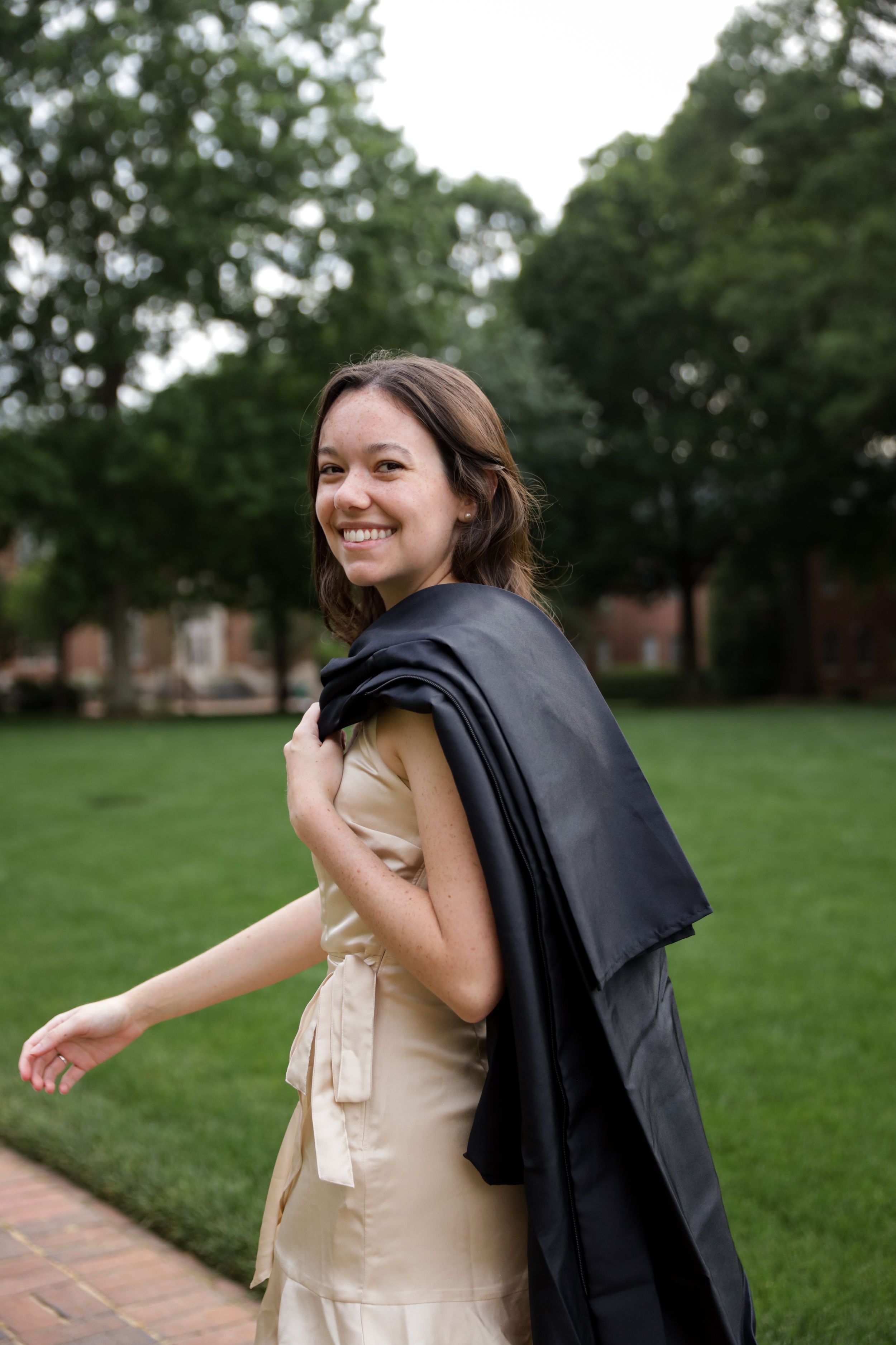 Joyful portrait of a graduate smiling at the camera, with her gown draped over her shoulder, in natural light on Davidson College's campus