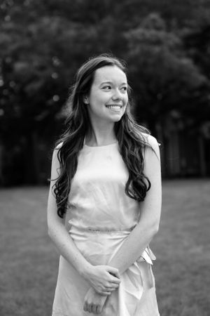 Artistic natural light black and white graduation portrait of a girl standing on on campus, smiling away from the camera.