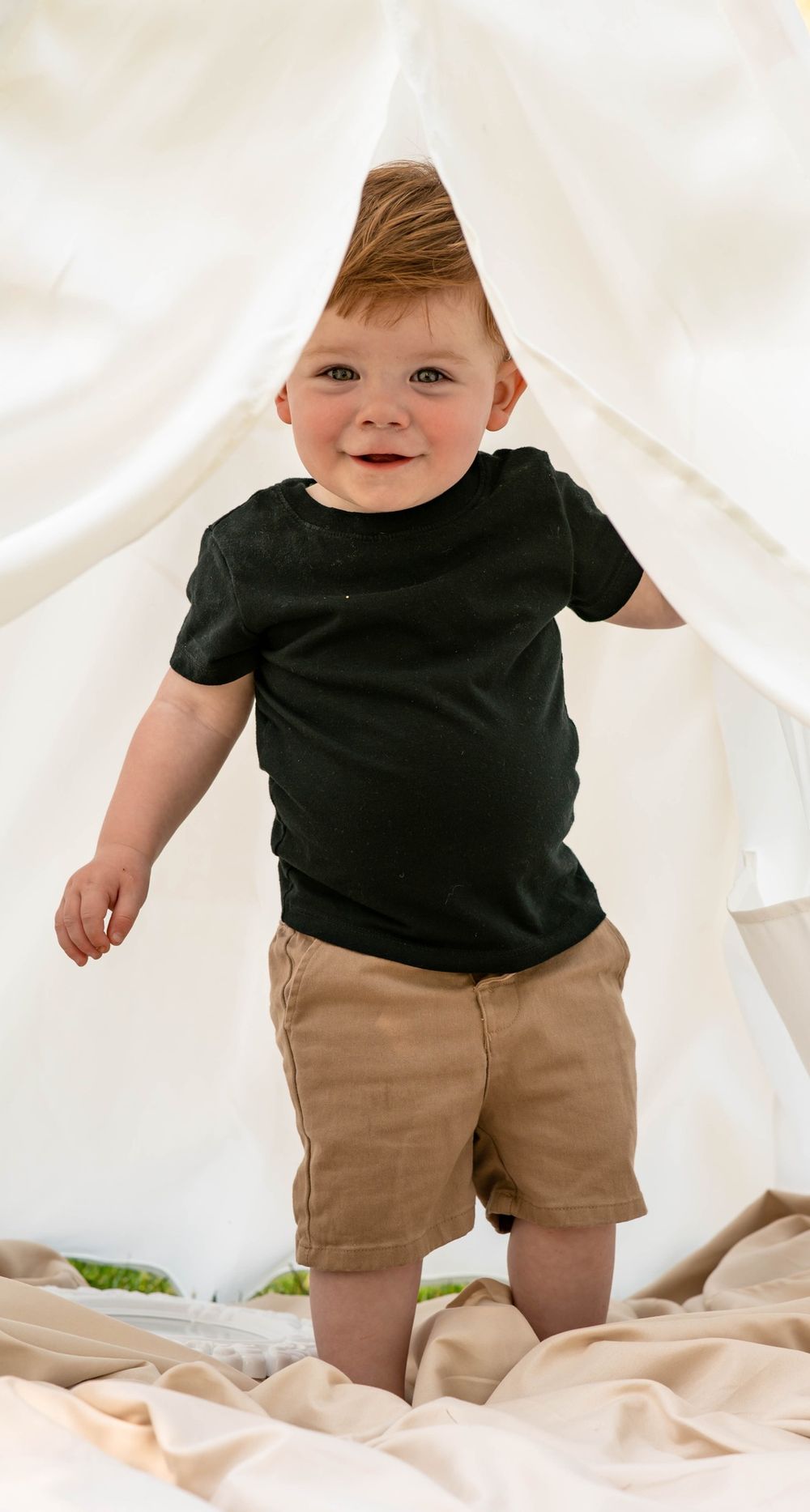 Smiling red-haired toddler boy in black shirt and tan shorts standing in white tent.