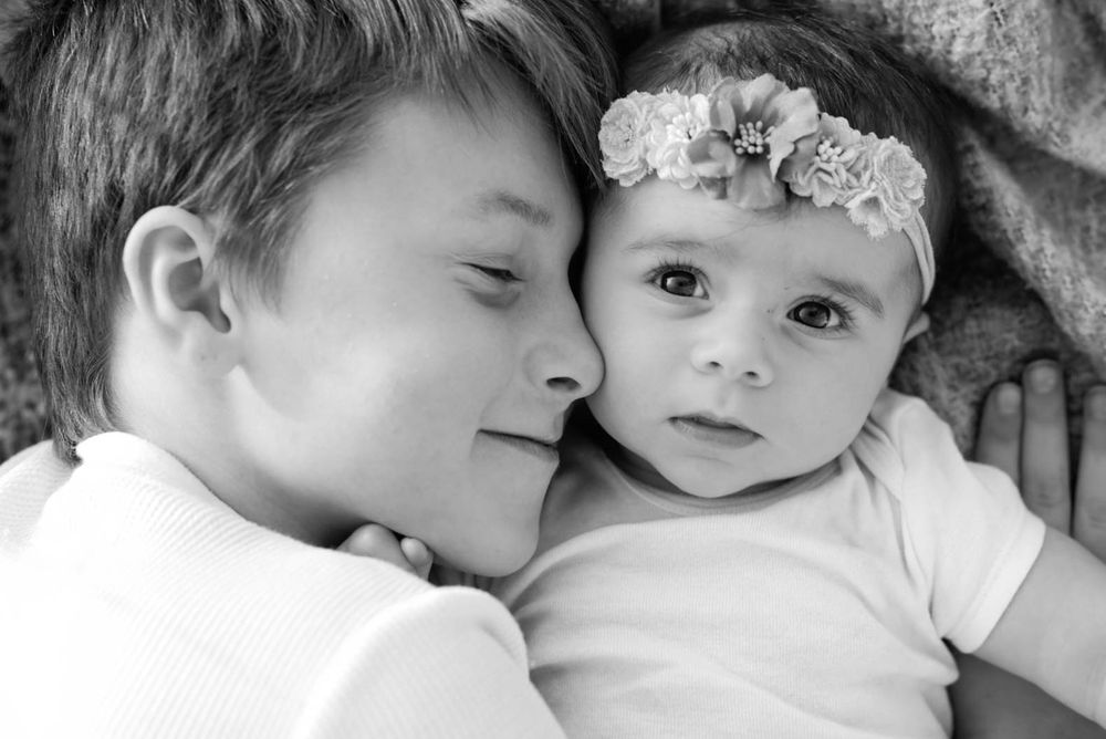 Black and white portrait of older brother cuddling newborn sister with floral headband.