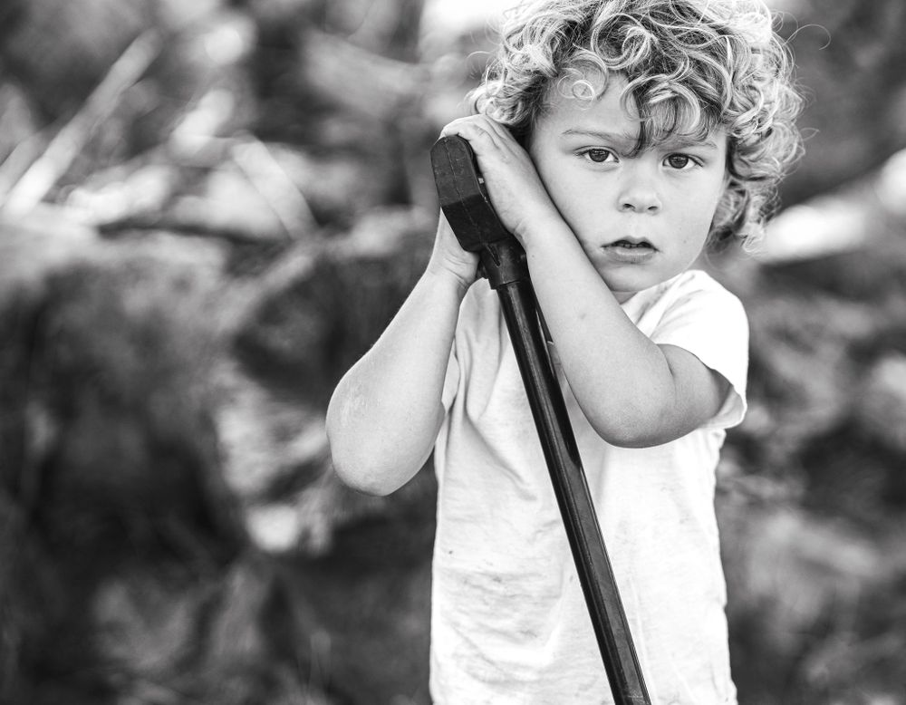 Black and white photo of a curly-haired boy leaning on a wooden stick outdoors.