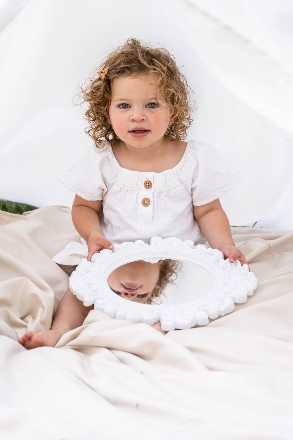 Curly-haired toddler girl in white dress holding an ornate mirror while sitting on fabric.